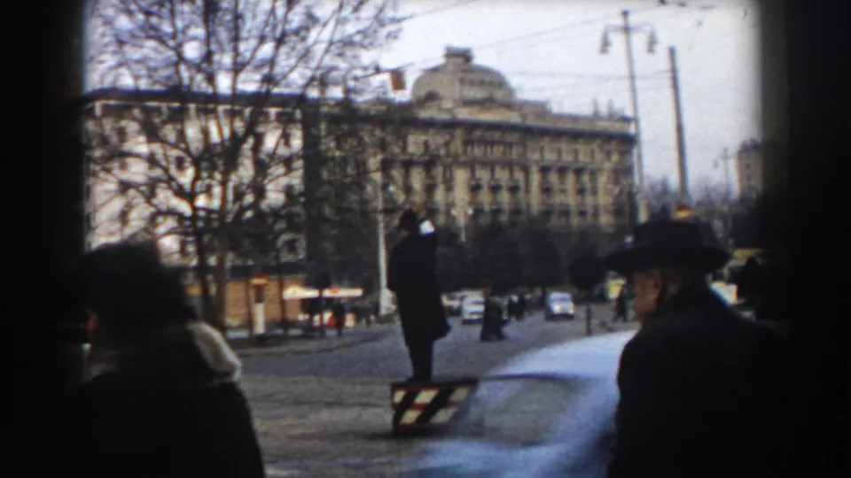 1959 8mm frame from Pisa with motion-blurred car, traffic officer, pedestrians and winter trees in a formal street setting.