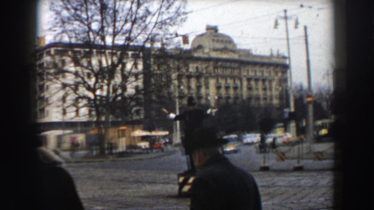 1959 Pisa street view with a traffic officer in the roadway, pedestrians nearby, compact cars, bare trees, overhead wires, and an ornate building backdrop.