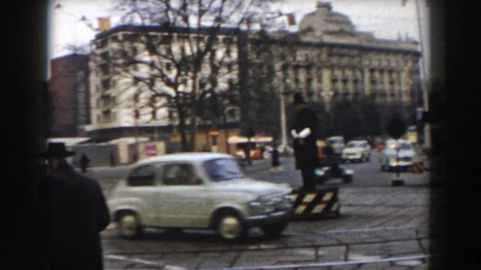 1959 Pisa traffic view with a light compact car crossing the foreground, a traffic officer standing beyond it, and pedestrians near an ornate building.