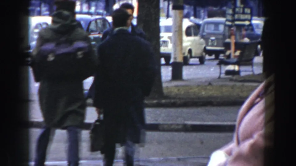 Two coat-wearing pedestrians seen from behind on a Pisa sidewalk, with compact cars and street furniture ahead.