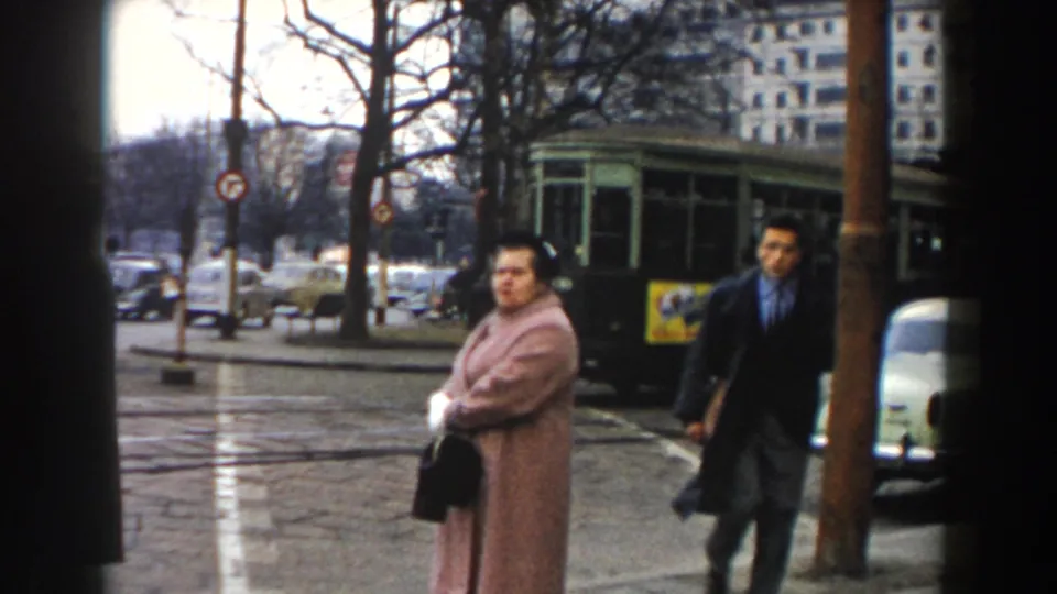 1959 Pisa street corner with a woman in a pink coat, a brown pole, a suited man, cars, trees, and a green kiosk or shelter.