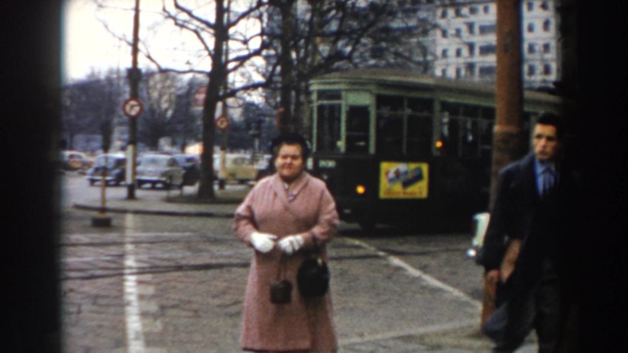 In 1959 Pisa, a woman in a pink coat faces the camera on a wet street with a green tram, cars, bare trees, and buildings behind her.