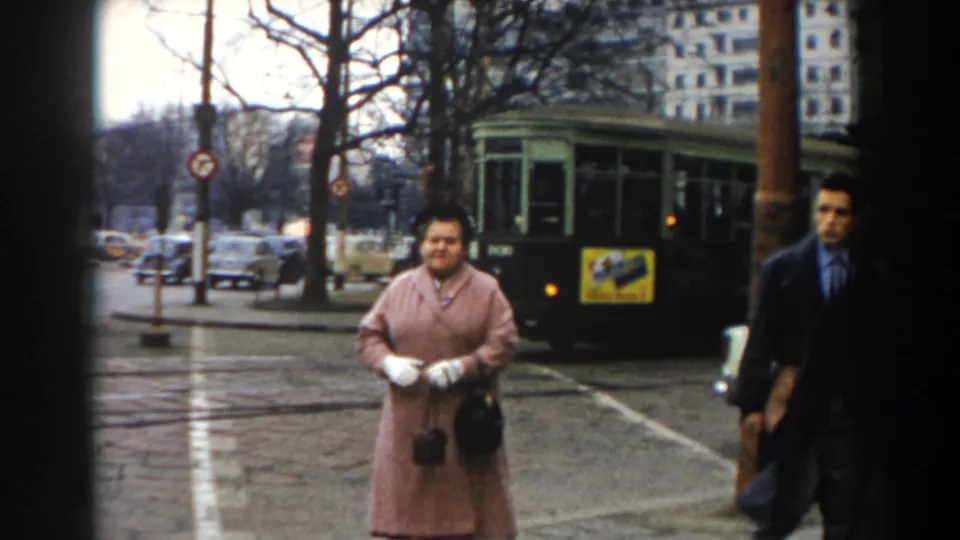 In 1959 Pisa, a woman in a pink coat faces the camera on a wet street with a green tram, cars, bare trees, and buildings behind her.