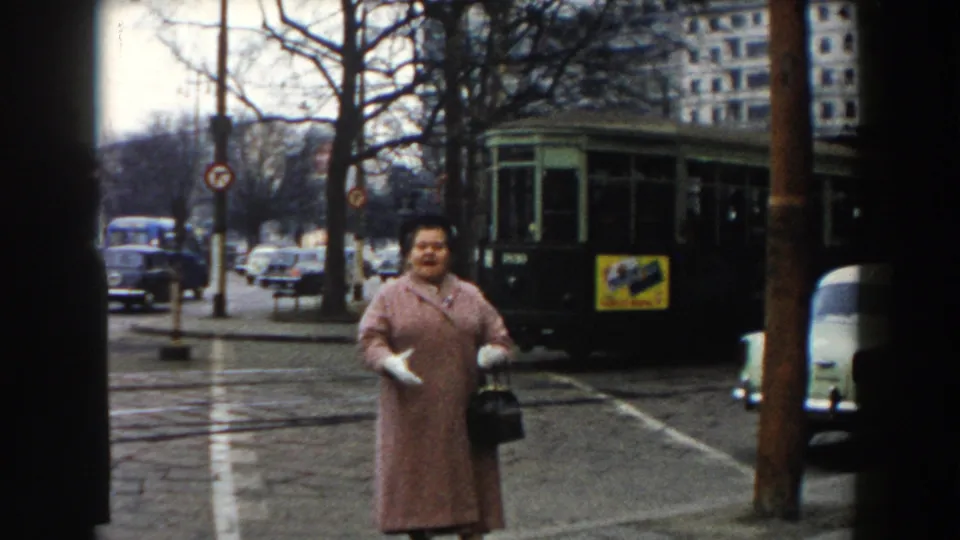 The woman in the pink coat gestures toward the camera on a Pisa street while a green tram and period cars pass behind her.
