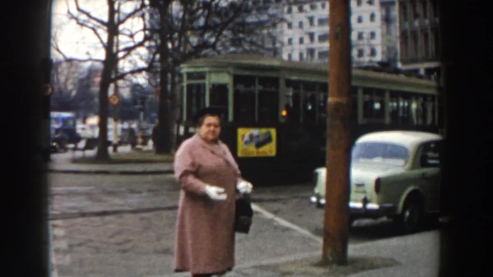 On the 1959 Pisa street, the woman turns in profile near a pole, holding a dark handbag as the tram and a pale compact car sit behind her.