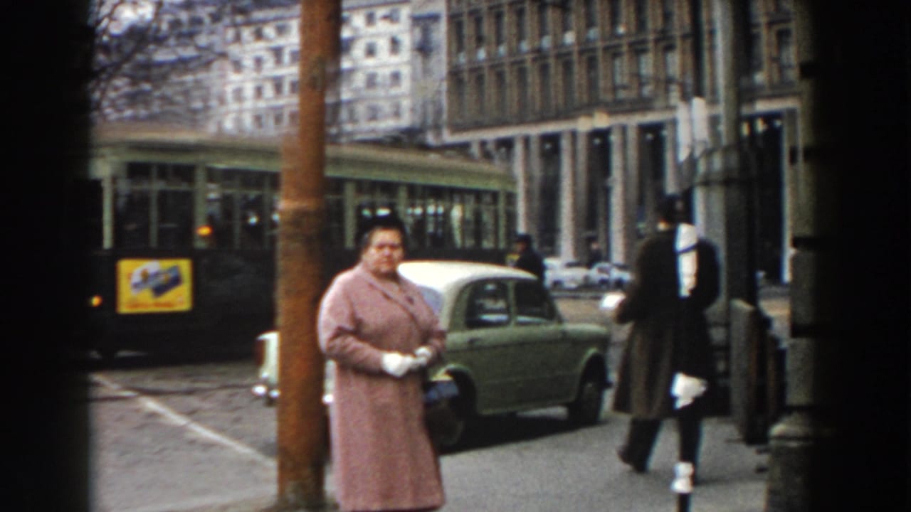 Woman in a pink coat stands by a street with a green car and tram behind her.