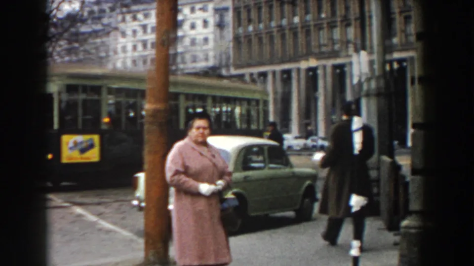 In Pisa, Italy, a woman in a pink coat and white gloves waits near the curb as a green car and tram pass behind her.