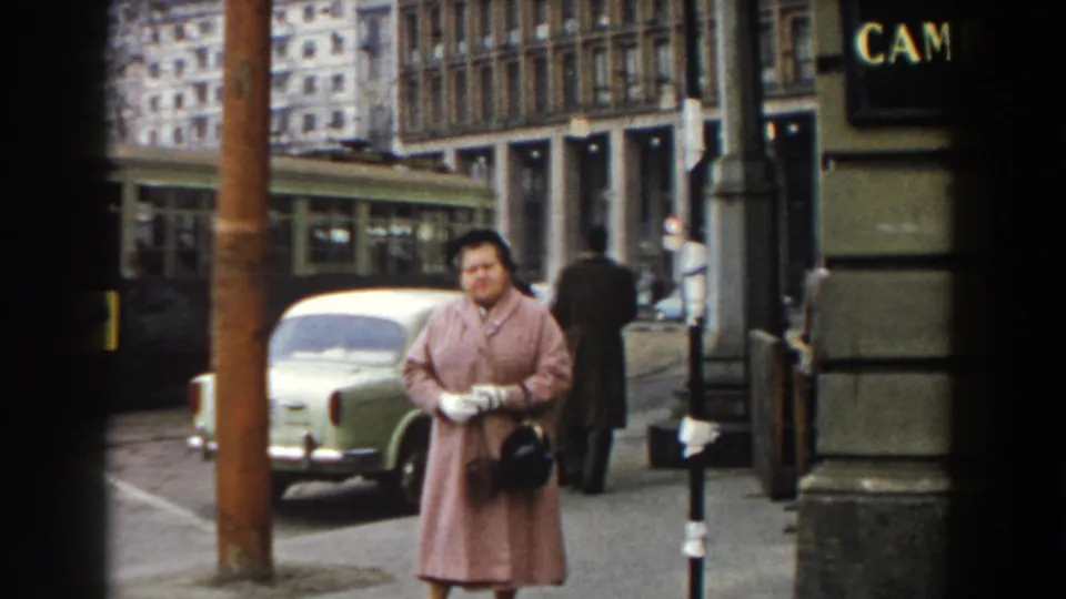 The woman in a pink coat stands near the curb in Pisa as a pale green car and pedestrians occupy the street behind her.