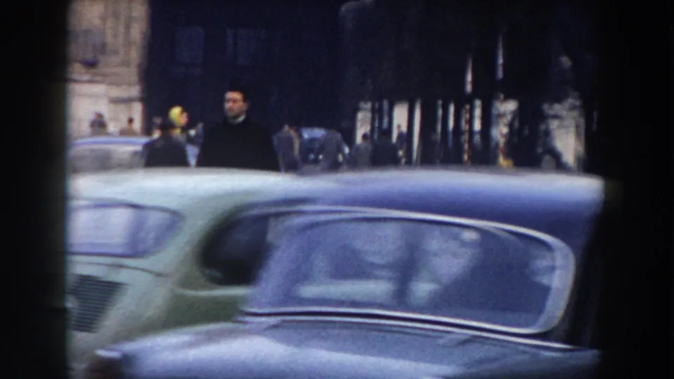 A vintage green car fills the foreground while pedestrians walk along a shaded Pisa street in the supplied 1959 home-movie footage.