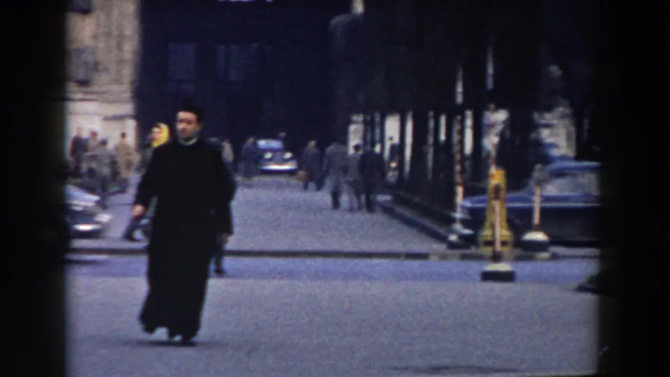 A man in dark clothing crosses a Pisa street while other pedestrians and parked or moving cars appear in the background.