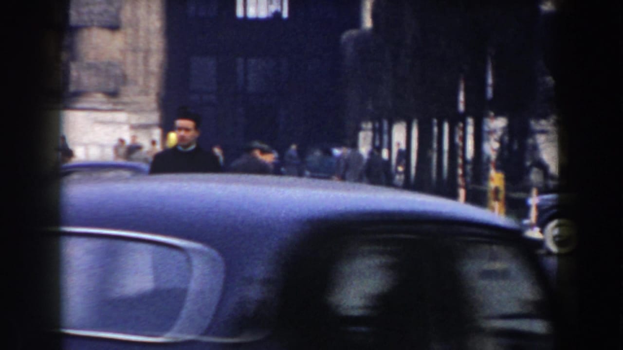 In Pisa, Italy, a clergyman in black is partly obscured by a dark vintage car passing across a busy 1959 street scene.