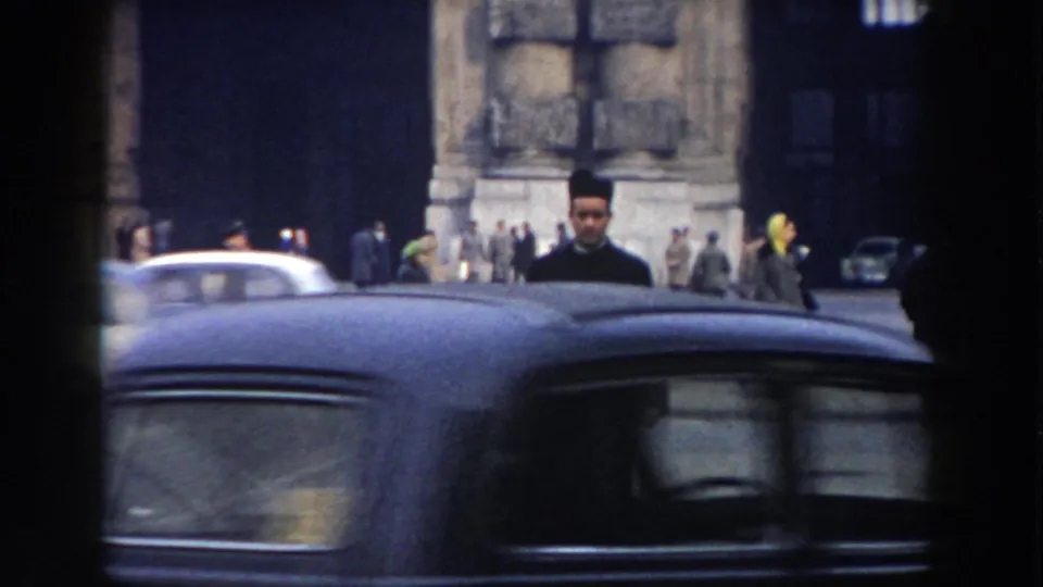 A black-robed clergyman stands or walks behind a passing dark car on a 1959 Pisa street with pedestrians in the background.