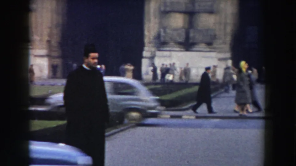 A clergyman in black walks through a Pisa street scene, with vintage cars, pedestrians, stone architecture, and a grass verge visible.