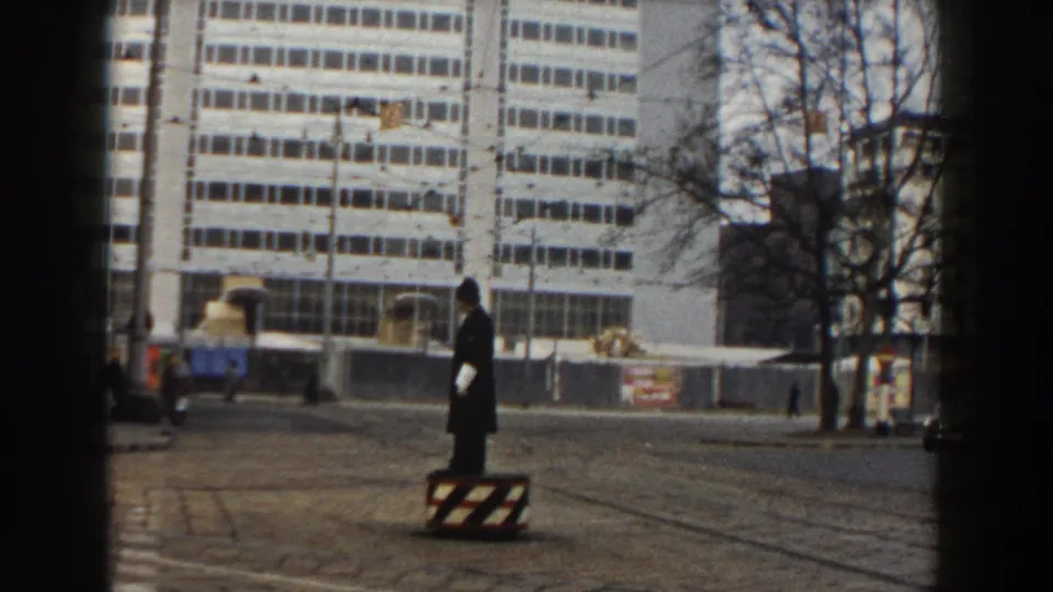 A Pisa traffic officer stands on a striped safety barrel while directing street traffic near a modern building and bare trees.