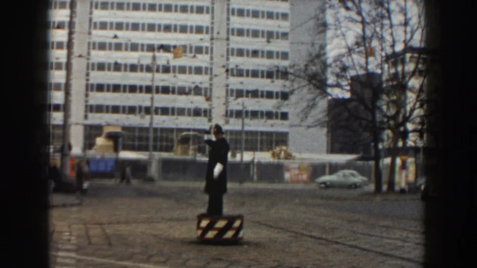The traffic officer continues directing movement from a striped stand as mid-century cars and pedestrians pass through the Pisa street scene.