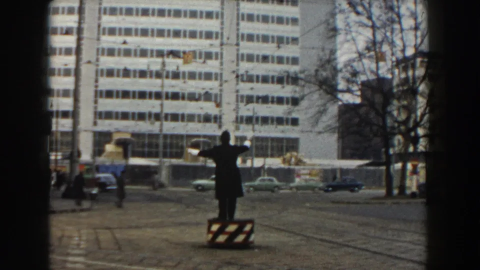 A uniformed traffic officer gestures from a striped stand in 1959 Pisa, with cars, pedestrians, trees, and a modern building behind him.