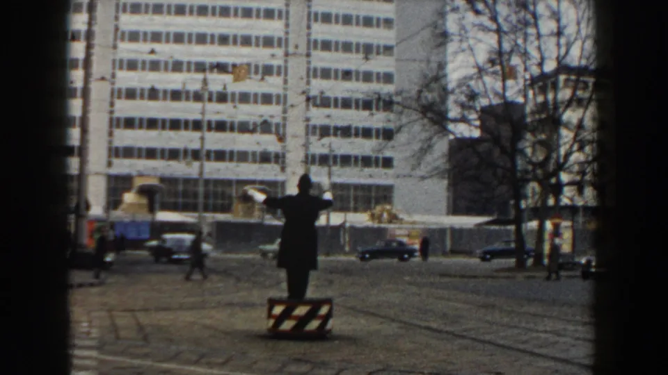 1959 Pisa street view with a dark-coated traffic director on a raised platform, tram rails, vintage cars, bare trees, and a large white building.