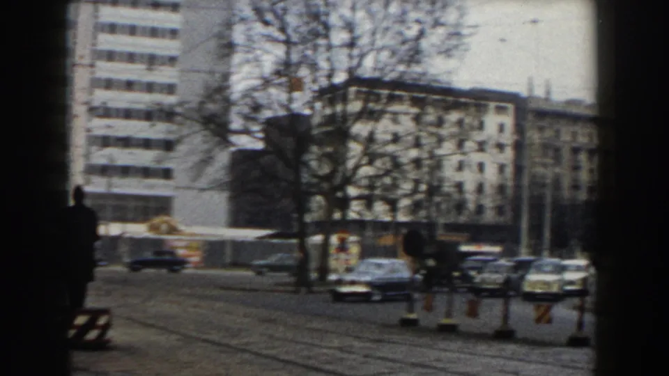 Pisa street scene from 1959 showing a traffic director in silhouette near tram rails, with cars, barriers, bare trees, and city buildings beyond.