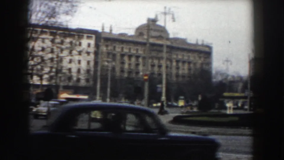 1959 Pisa street view with a dark car passing in the foreground, a planted traffic island, bare trees, street poles, and masonry buildings.