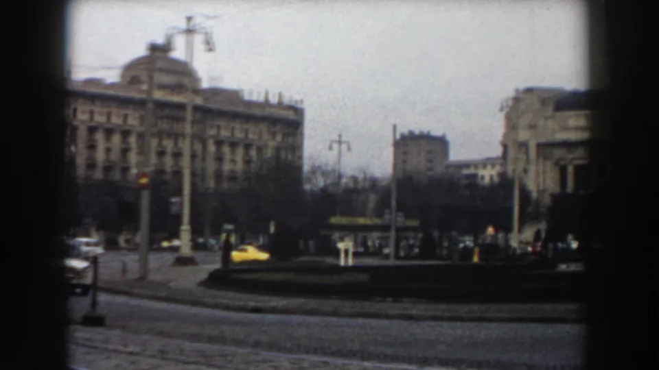 Pisa street view from 1959 with a yellow car near a traffic island, pedestrians at distance, bare trees, street poles, and surrounding buildings.