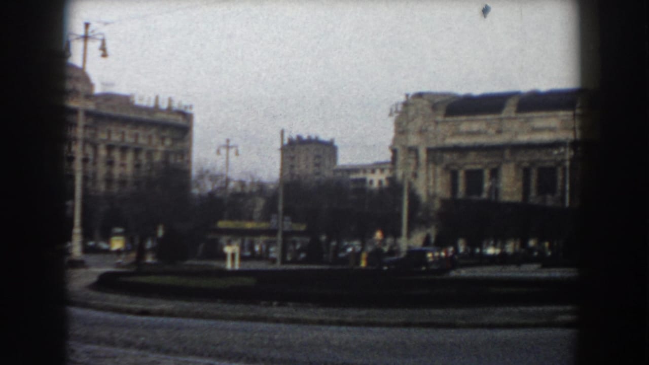 1959 Pisa street view with masonry buildings, a traffic circle, cars, poles, overhead cables, and distant pedestrians framed by dark window edges.