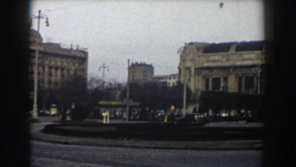1959 Pisa street view with masonry buildings, a traffic circle, cars, poles, overhead cables, and distant pedestrians framed by dark window edges.