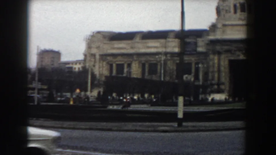 1959 Pisa city scene showing a large stone building, circular roadway, passing car, street poles, and small groups of distant pedestrians.