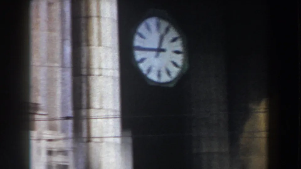 1959 Pisa 8mm frame with a large clock face, dark wall surface, pale stone columns, and railings visible in a blurred passing view.