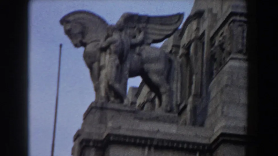 1959 8mm frame identified as Pisa, Italy, showing a winged sculpted figure on a historic stone facade against a pale sky.