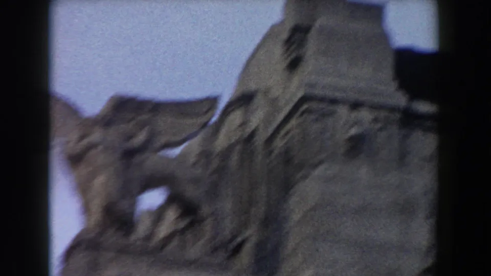 Soft 8mm view of a winged stone sculpture and rounded facade detail, partially blurred by camera movement and dark frame edges.