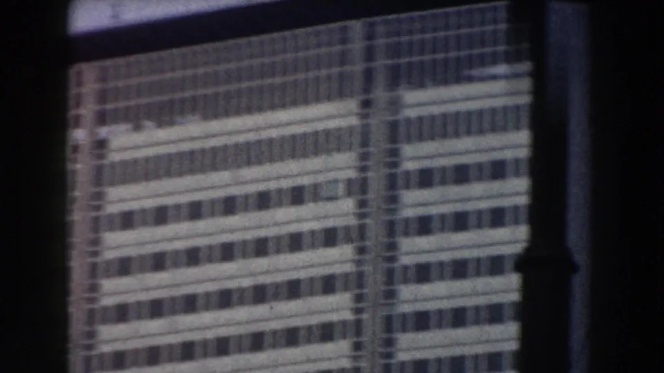 Handheld 8mm frame from Pisa showing a gridded modern facade partly crossed by shadowed foreground structure.