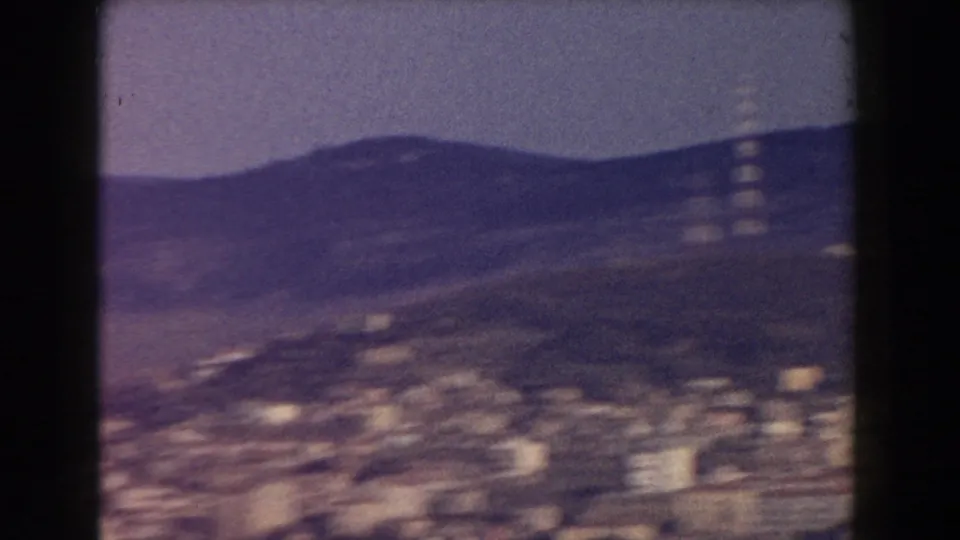 1959 Pisa aerial footage frame with blurred pale buildings at the base of hills, framed by dark 8mm contact-sheet borders.