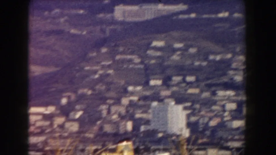 1959 Pisa home-movie frame looking down over distant urban blocks, pale buildings, hillside terrain, and soft 8mm grain.