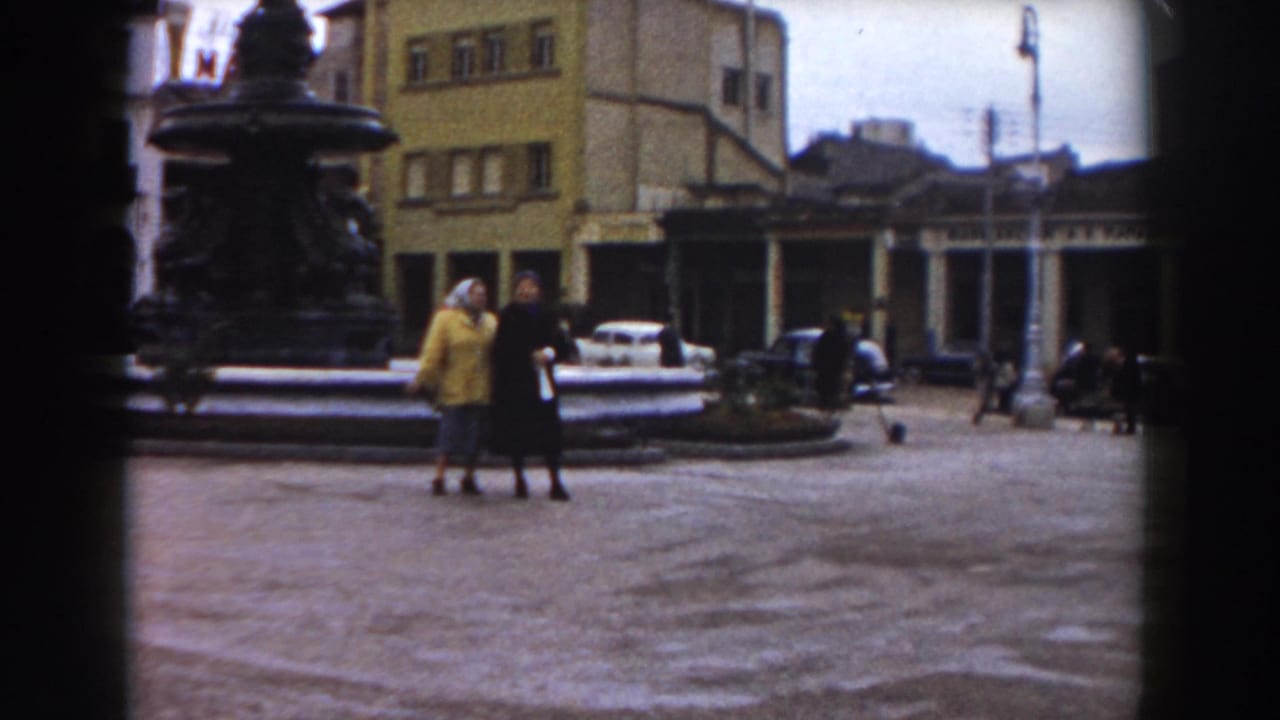Two women walk in a paved square beside a large fountain, with buildings and cars behind them.