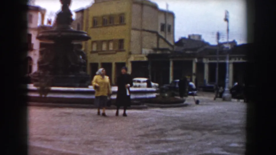 The women continue across the Pisa square, framed by the fountain, parked cars, street lamps, and mid-century buildings.