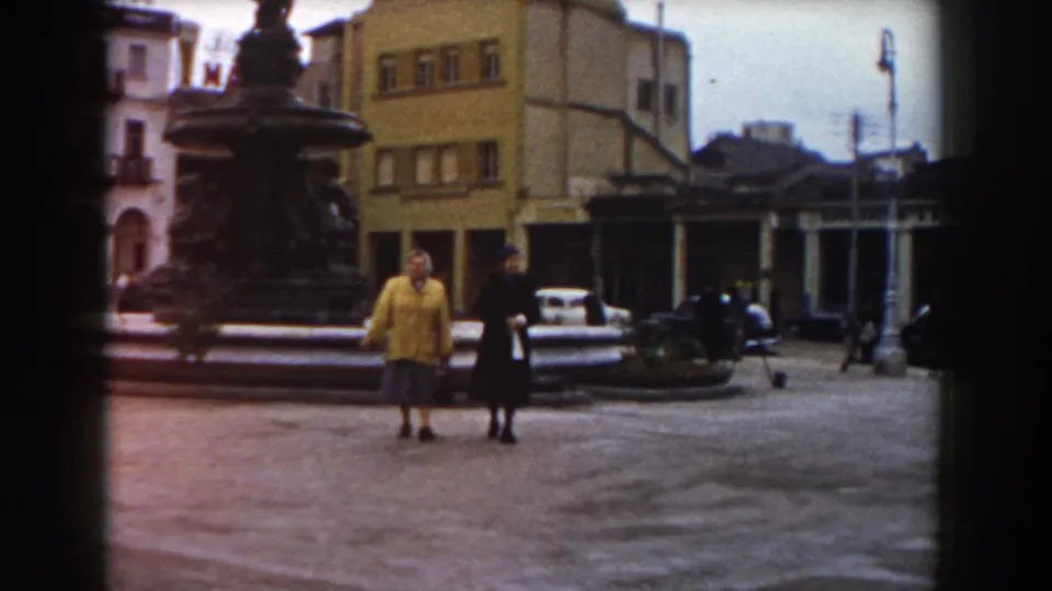 In the 1959 Pisa home-movie frame, the women pause or slow near the fountain, with a white car and square-front buildings behind them.