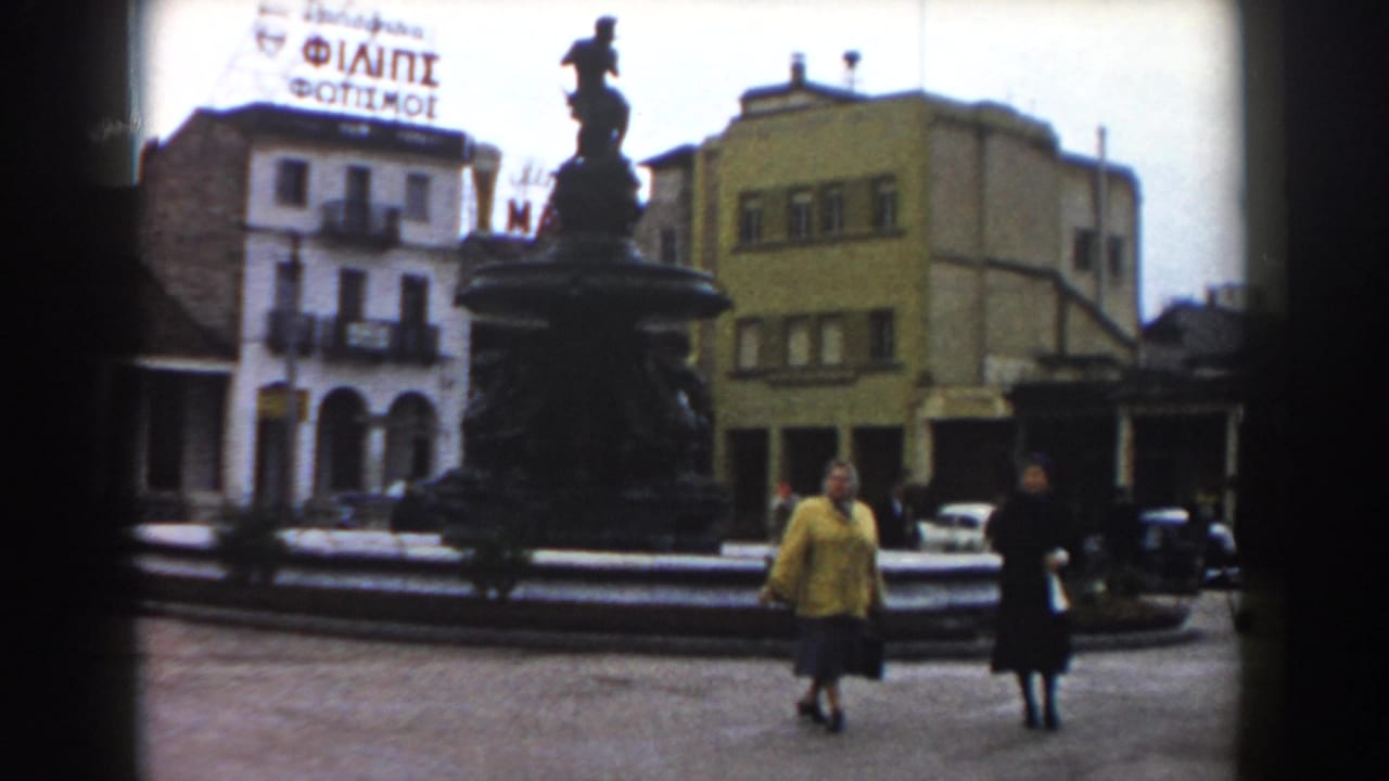 Pisa, Italy, 1959 home-movie frame showing two women crossing a square near an ornate fountain, with arched buildings and signage in the background.