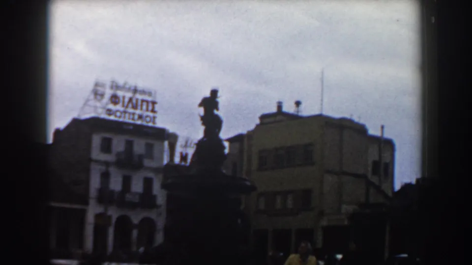 Pisa, Italy, 1959 frame with the fountain in silhouette, surrounding mid-century buildings, a rooftop sign, and partial pedestrians at the lower edge.