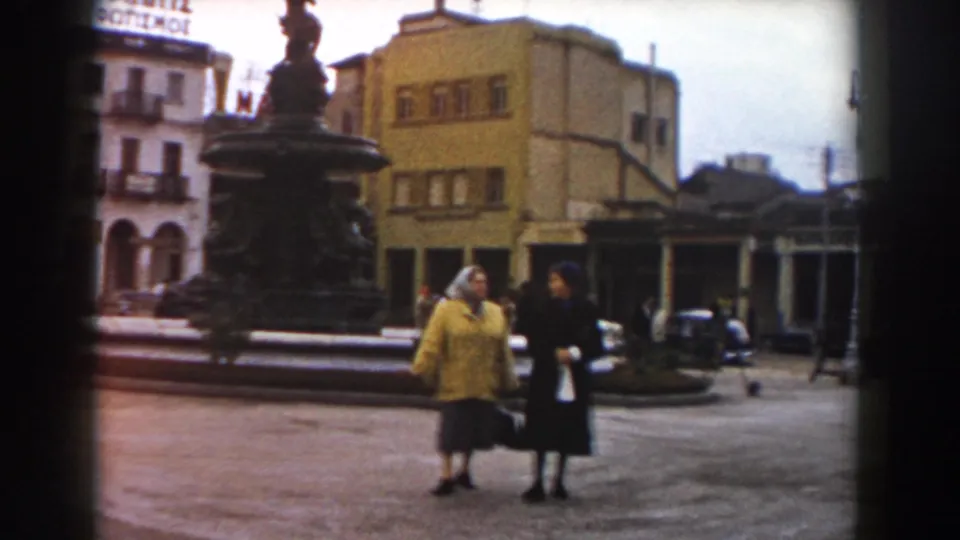 Pisa, Italy, 1959 frame of two women near the fountain in a public square, with parked cars, arcaded buildings, and a visible rooftop sign behind them.