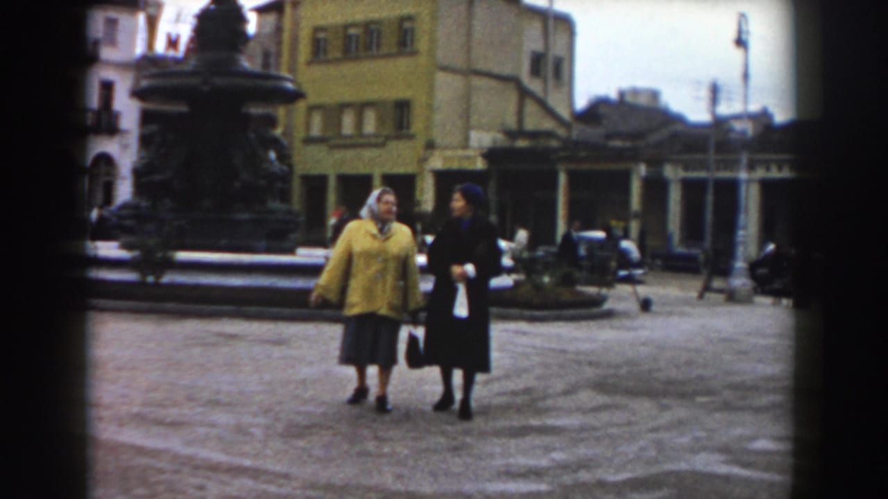 Two women walk across a town square near a large dark fountain.