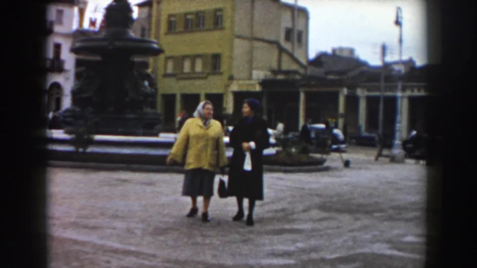 In Pisa, Italy, 1959, two women walk and talk in a public square near an ornate fountain, with buildings and parked cars behind them.