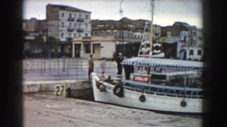 A white ferry named ITALIA sits at pier 27 in a Pisa harbor scene, with people on the dock and waterfront buildings in the background.