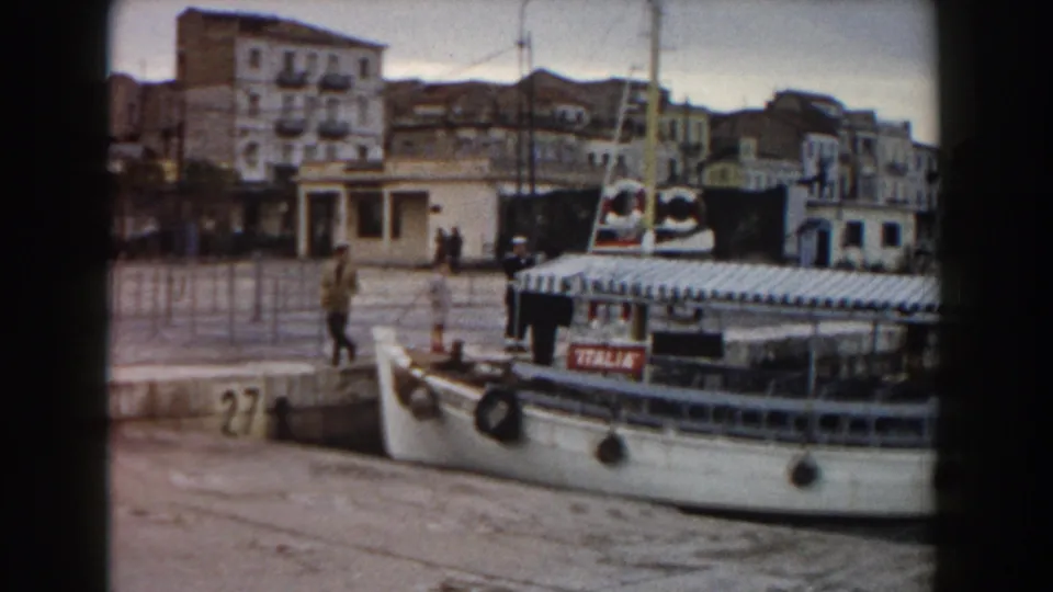 The docked ITALIA ferry fills the foreground at pier 27, while a few people stand or walk along the quay in front of Italian waterfront buildings.
