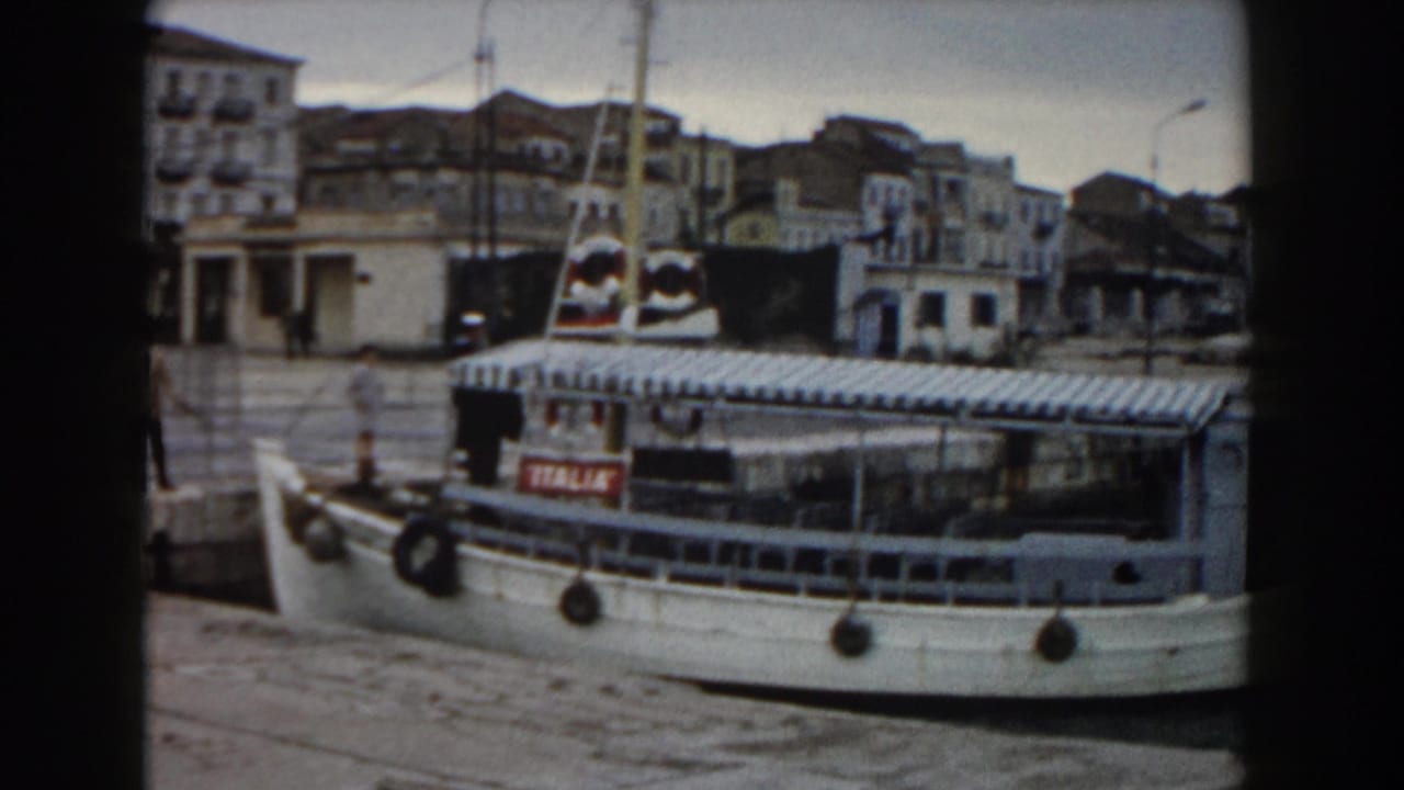 Small white ferry beside a stone pier with waterfront buildings behind it.