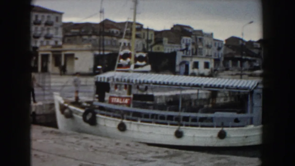 1959 Pisa harbor view of a small white ferry with a striped canopy docking near a stone pier, with buildings in the background.