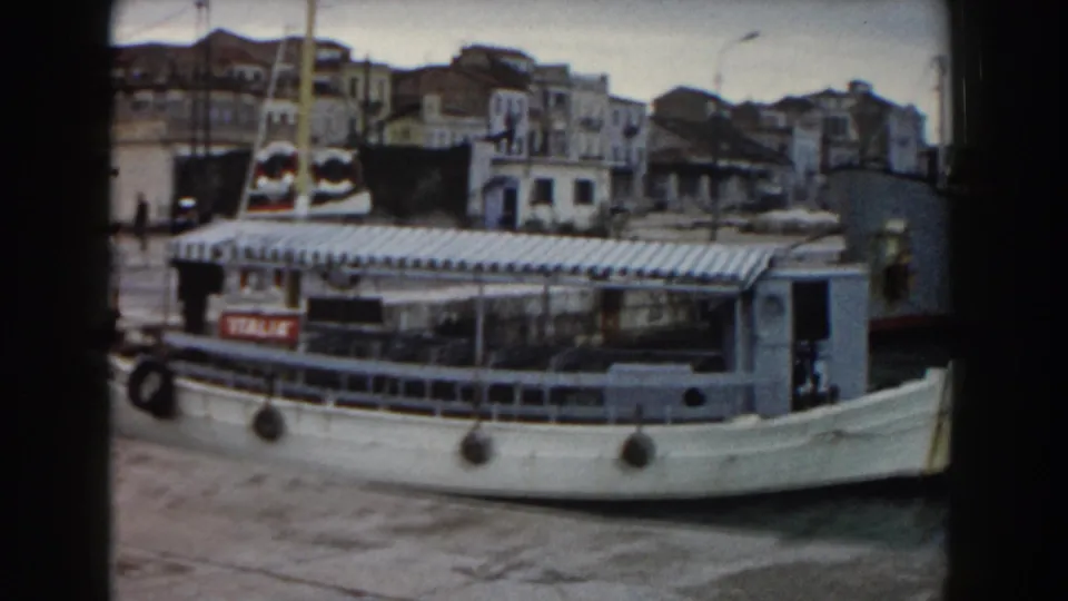The white ferry remains alongside the Pisa harbor quay, showing its canopy, side rail, fenders, and the waterfront beyond.