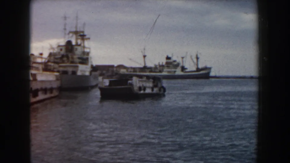 1959 Pisa harbor view with a small boat in front of larger ships, cranes or masts, open water, and cloudy sky.