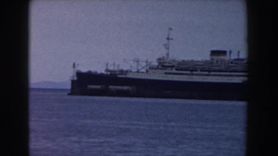 The docked liner becomes clearer across the water, with a dark hull, white upper decks, mast and funnel visible.