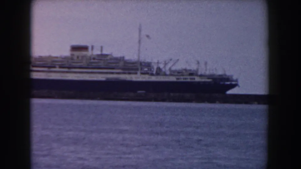 The bow and forward decks of the docked liner are visible across the harbor, with masts and dock equipment silhouetted nearby.