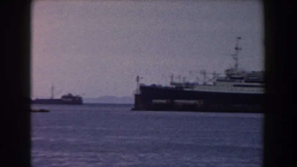 The camera frames the ship partly at right, with open harbor water and a smaller vessel or structure visible in the distance.